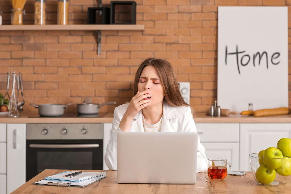 Une femme fatiguée bâille devant un ordinateur portable dans une cuisine.